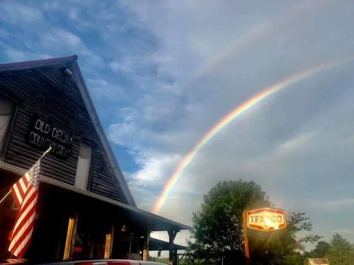 A rustic building with an American flag, under a sky featuring a double rainbow and a Texaco sign.