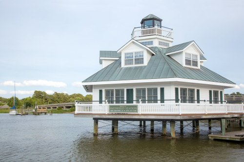 A white waterfront building with a green roof, sitting on stilts over calm water, surrounded by trees and a boat dock.