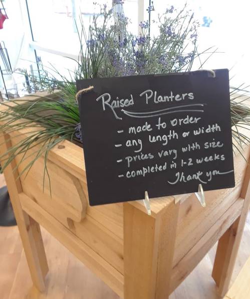 A wooden raised planter display with a chalkboard sign detailing custom orders and pricing information.