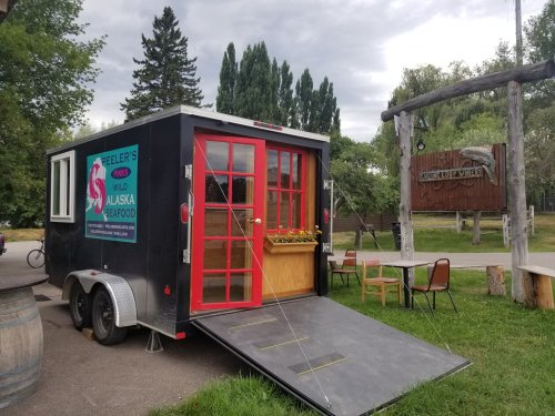 A food truck with red doors and a wooden serving window, parked near a sign for "Hooligan Park" surrounded by trees.