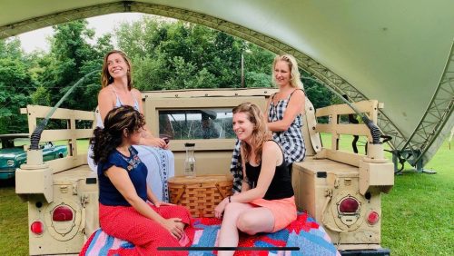 Four women sitting on a vintage vehicle, smiling and enjoying each other's company under a large canopy.