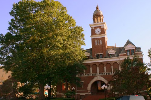 Historic brick building with a clock tower, surrounded by trees and clear blue sky.