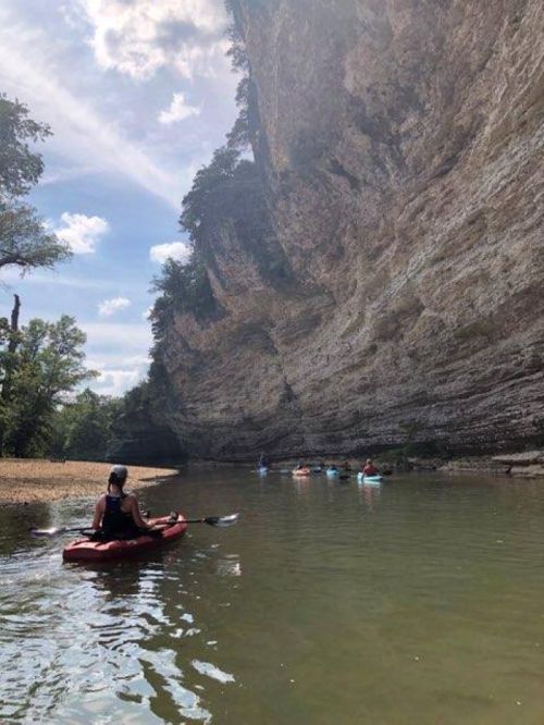 A person kayaking in a calm river surrounded by steep, rocky cliffs and lush greenery under a blue sky.