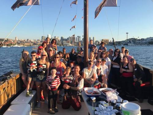 A large group of people in festive attire poses on a boat with a city skyline in the background on a sunny day.