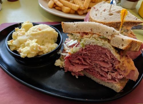 A large sandwich stacked with layers of meat and toppings, served with a side of potato salad on a black plate.