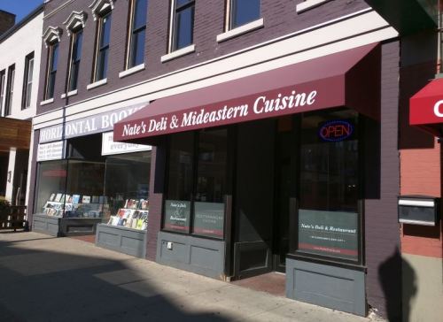 Exterior of Nate's Deli & Mideastern Cuisine with a red awning and "OPEN" sign, next to a bookstore.