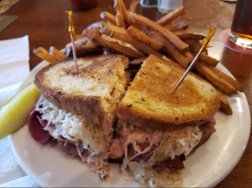 A sandwich with corned beef and sauerkraut, served with golden fries and a pickle on a white plate.