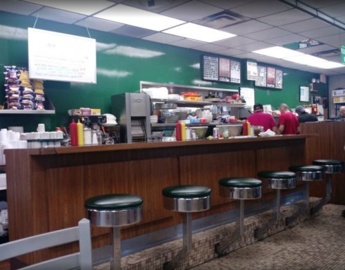 A diner interior featuring a wooden counter, green walls, and bar stools, with staff preparing food in the background.