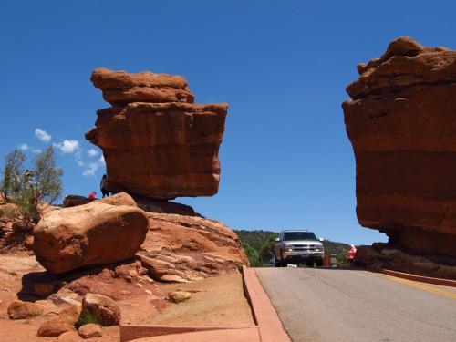 A car drives through a narrow passage between two large rock formations under a clear blue sky.