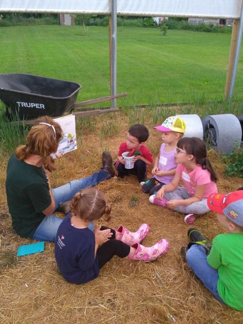 A group of children sitting on straw, listening to an adult in a garden setting, with tools and greenery in the background.