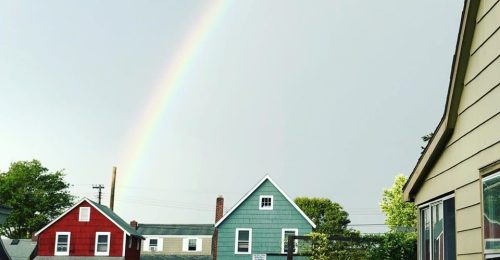 A vibrant rainbow arcs over colorful houses under a cloudy sky.