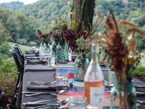 A beautifully set outdoor dining table with flowers in jars, surrounded by greenery and a scenic view.