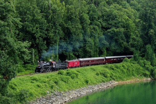 A vintage steam train travels along a lush green forested area beside a calm body of water.