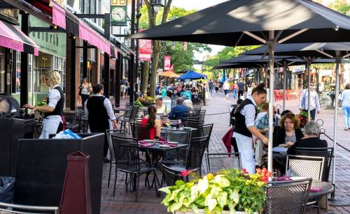 Outdoor dining scene with people at tables, waitstaff serving, and shops in a bustling street.