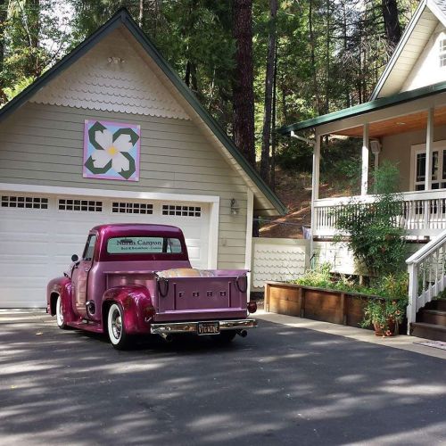 A vintage purple truck parked in front of a house with a decorative garage door and lush greenery.