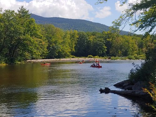 A serene river scene with kayakers paddling, surrounded by lush green trees and mountains under a blue sky.