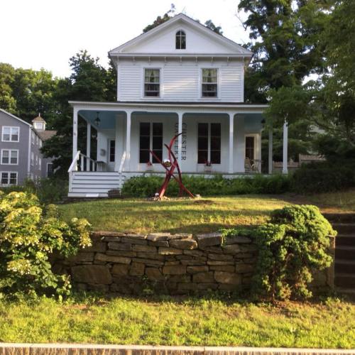 A white two-story house with a porch, surrounded by greenery and a stone wall, featuring a red sculpture in the yard.