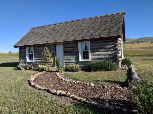 A rustic log cabin with a sloped roof, surrounded by a landscaped garden and grassy fields under a clear blue sky.