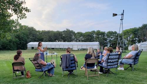 A group of women sitting in chairs outdoors, listening to a speaker near a greenhouse and a windmill.