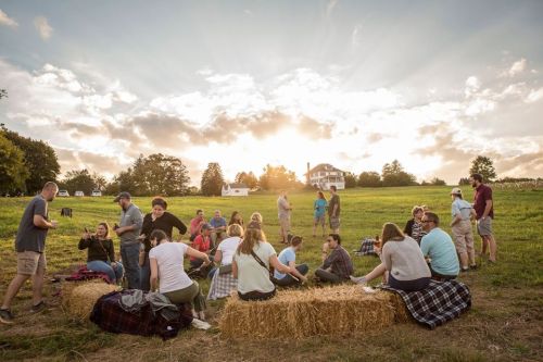 A group of people gathered on hay bales in a field during sunset, enjoying a social event with a house in the background.