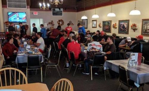 A busy restaurant scene with many people seated at tables, some watching a TV, and autumn decorations on the walls.