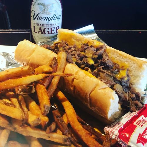 A plate featuring a cheesesteak sandwich with fries and a bottle of Yuengling lager.