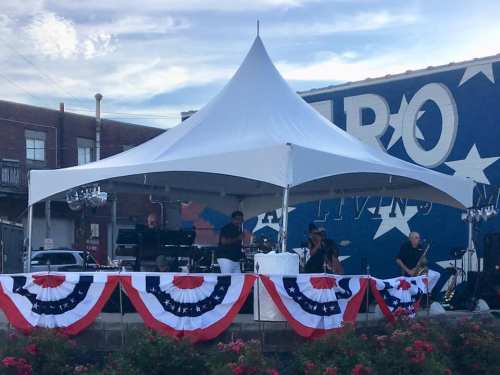A band performs under a large tent decorated with red, white, and blue bunting, with a mural in the background.