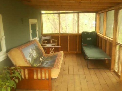 Cozy screened porch with a wooden floor, a futon, a chair, and a folding cot surrounded by trees.