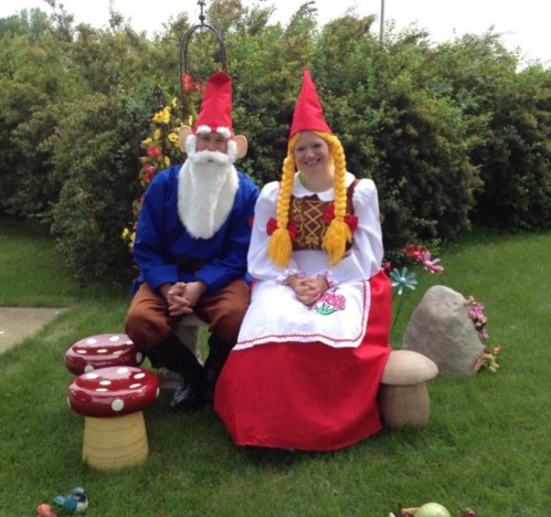 A man with a white beard and red hat sits beside a woman in a red dress and braids, both in a garden setting.