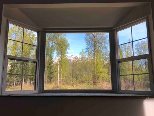 A scenic view from a window, showcasing trees and a snow-capped mountain under a clear blue sky.