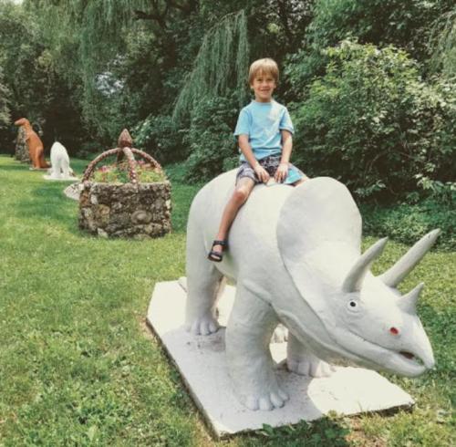 A child sits on a white dinosaur statue in a grassy area, with other colorful dinosaur sculptures in the background.