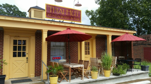 Exterior of Thai Hut restaurant with a sign, outdoor seating, and red umbrellas on a sunny day.