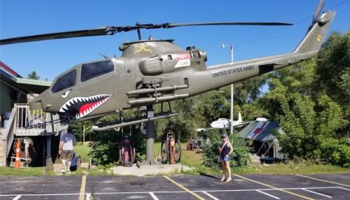 A military helicopter on display, with a shark mouth design, near a building and visitors in a parking lot.