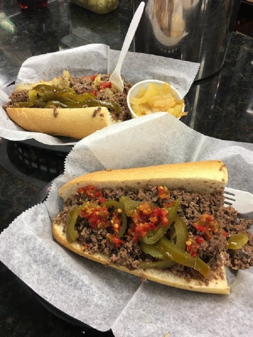Two sandwiches filled with seasoned ground beef, topped with green peppers and spicy sauce, served with a side of pickles.