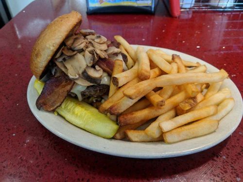 A hearty burger topped with mushrooms and bacon, served with crispy fries and a pickle on a plate.