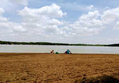 Two people sit on a sandy beach by a calm lake under a partly cloudy sky. Green trees line the far shore.