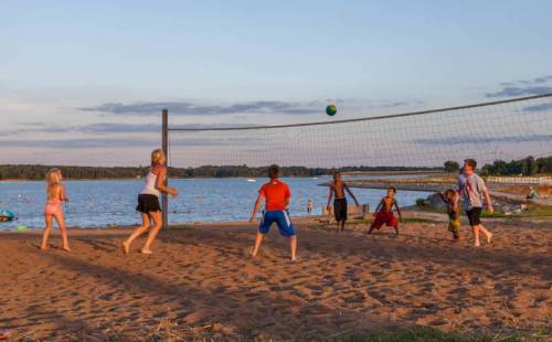 Children playing beach volleyball near a lake during sunset, with a sandy shore and water in the background.