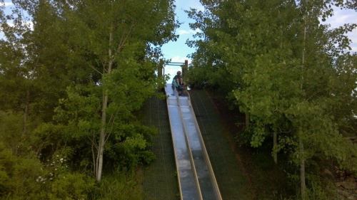A metal slide on a grassy hill surrounded by trees, with two people sitting at the top.