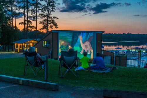 People sitting on the grass watching a movie projected on a screen at sunset by a lake.