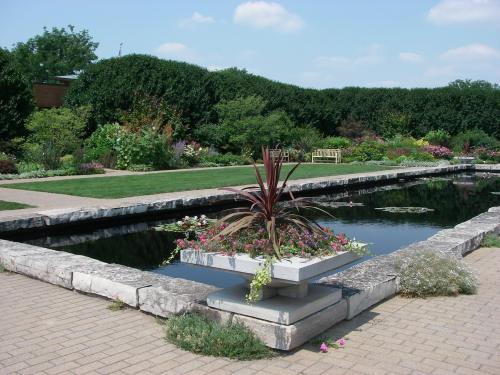 A serene garden with a reflecting pond, surrounded by lush greenery and colorful flowers under a clear blue sky.