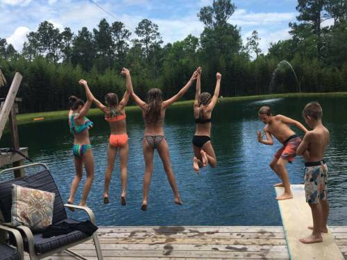 A group of children jumps off a dock into a lake on a sunny day, surrounded by trees and water.