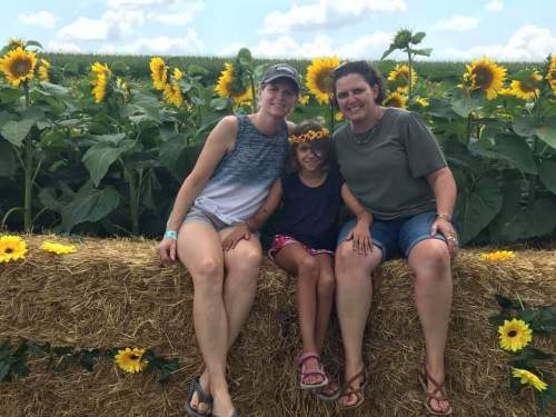 Three people sit on a hay bale surrounded by sunflowers, smiling under a blue sky with fluffy clouds.