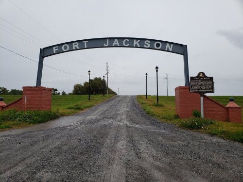 Archway sign reading "Fort Jackson" over a gravel road leading into a grassy area, with historical markers on the sides.