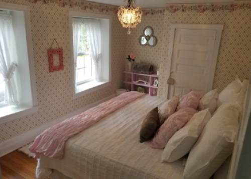 Cozy bedroom with a white bed, pink accents, floral wallpaper, and natural light from two windows.