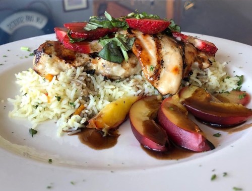 Grilled chicken atop rice, garnished with strawberries, basil, and served with sliced plums and a drizzle of sauce.