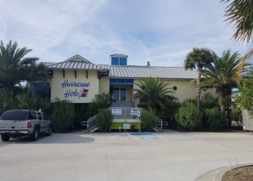 A yellow building with a metal roof, labeled "Hurricane Hole," surrounded by palm trees and parked vehicles.