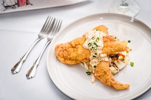 A plate of fried fish topped with creamy sauce and green onions, served on a bed of coleslaw. Forks in the background.