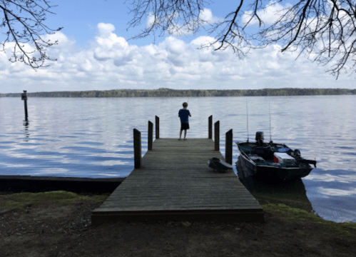 A child stands on a wooden dock by a calm lake, with a small boat nearby and trees in the background under a cloudy sky.
