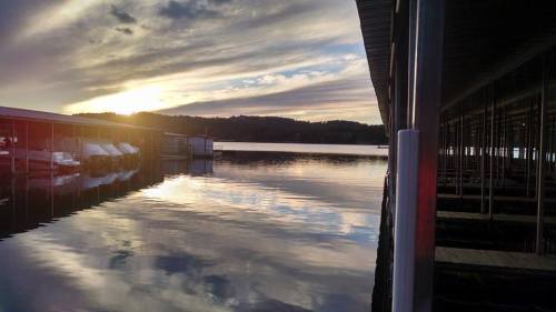 A serene lake at sunset, reflecting clouds and boats under a dock, with hills in the background.