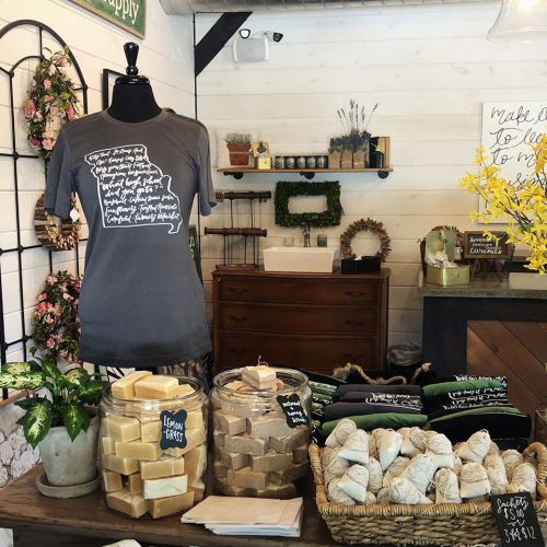A cozy shop display featuring a t-shirt on a mannequin, jars of soap, and decorative plants on a wooden table.
